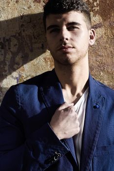 Close-up portrait of a young man with blue eyes posing near a wall. Model of fashion in urban background wearing white t-shirt and blue jacket