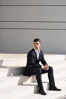 Young businessman near a modern office building wearing black suit and white shirt sitting on the floor. Man with blue eyes in urban background