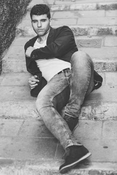 Good looking young man with blue eyes in the street. Model of fashion sitting in urban stairs wearing white t-shirt, jeans and blue jacket