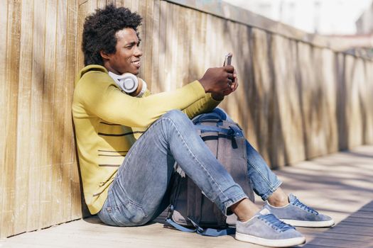 Black man with afro hair and headphones resting sitting on the ground using smartphone.