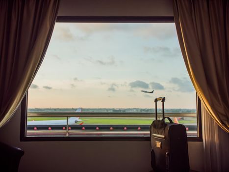 The window of the hotel near the airport and the plane taking off in the background of a suitcase or luggage