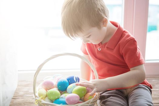 Little cute boy sits by the window with a full basket of colorful Easter eggs. .