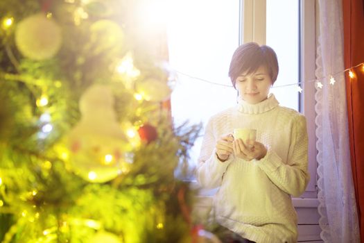 Happy young woman with cup of coffee or tea at home over christmas tree.