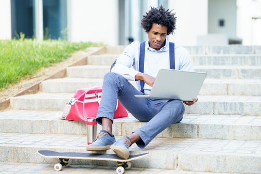 Black businessman with afro hair and skateboard using his laptop computer sitting on some steps of an office building.