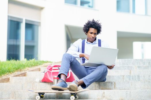 Black businessman with afro hair and skateboard using his laptop computer sitting on some steps of an office building.