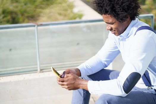 Black man with afro hairstyle using a smartphone near an office building. Guy with curly hair wearing shirt and suspenders.