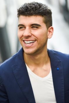 Good looking young man with blue eyes smiling in the street. Model of fashion in urban background wearing white t-shir and blue jacket