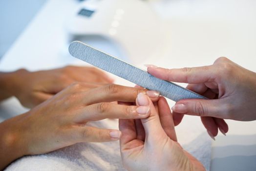 Close-up of a woman in a nail salon receiving a manicure by a beautician with. Beautician file nails to a customer.