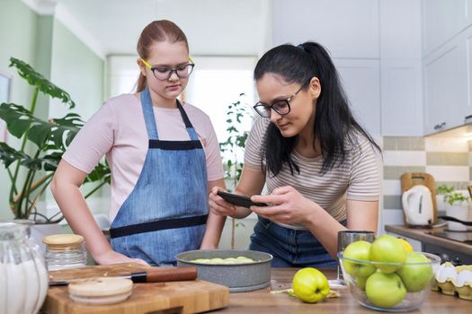 Mom and teenage daughter preparing apple pie together, at home in the kitchen. Woman photographing pie on smartphone. Family, parent teenager relationship, lifestyle, eating at home concept