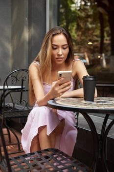 young well dressed woman sitting at table with drink, using her smartphone. lady having cup of coffee outside of cafe, surfing internet on cell phone, chatting online. modern communication technology