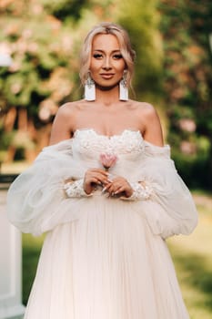 A beautiful bride in a luxurious wedding dress holds a rose and greenery on a green natural background. Portrait of a happy bride in a white dress, smiling against the background of a Park