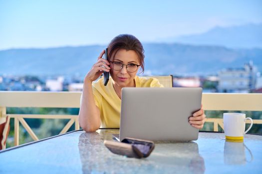 Middle aged woman working at home on terrace with laptop smartphone. Business female talking with client, colleague on phone, looking at laptop screen. Freelance, remote work, technology in business