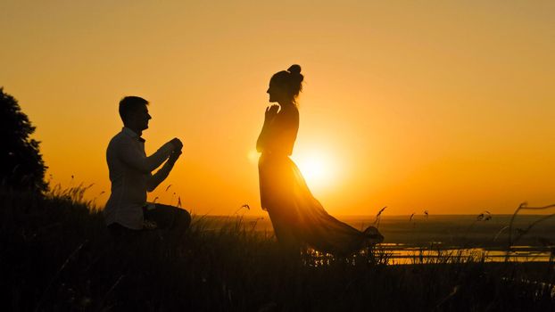 Romantic Silhouette of Man Getting Down on his Knee and Proposing to Woman on high hill - Couple Gets Engaged at Sunset - Man Putting Ring on Girl's Finger, cslider shot, medium angle
