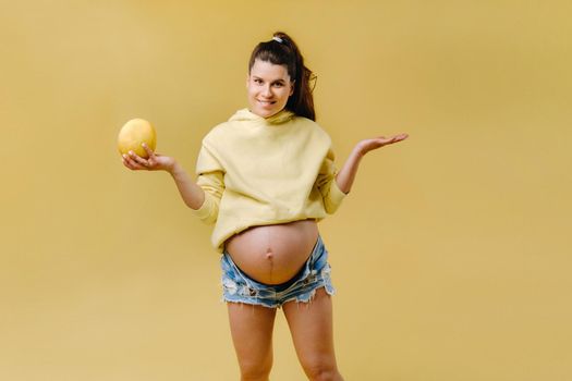 a pregnant girl with a yellow jacket stands with a melon in her hands on a yellow isolated background.