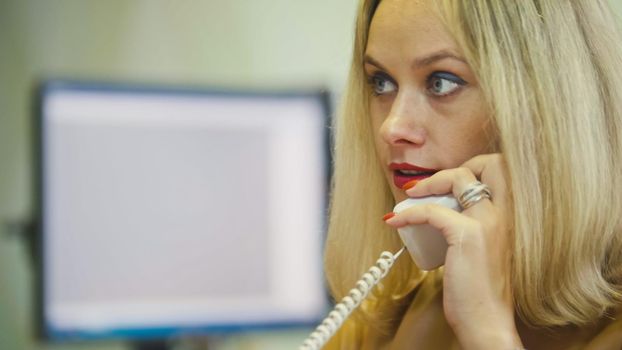 Young Blonde Woman in office talking on phone in front of the computer, close up, telephoto