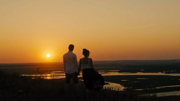 loving couple - brave young man and beautiful girl stands on high hill and looking to sunset silhouette, wide angle