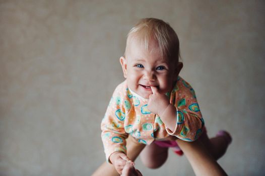 A little girl in her mother's arms above her head smiles with a finger in her mouth.