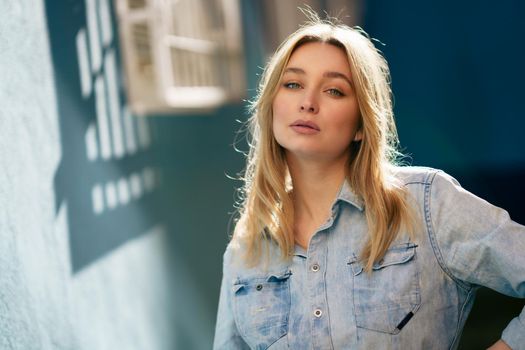 Portrait of blonde woman wearing denim shirt standing in the street. Pretty russian female in urban background.