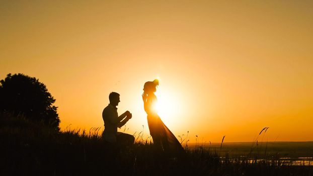 Silhouette of Man Getting Down on his Knee and Proposing to Woman high hill - Couple Gets Engaged at Sunset - Putting Ring Girl's Finger, telephoto