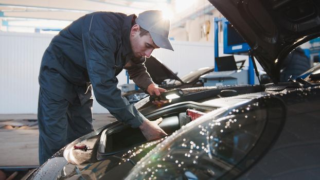 Mechanic male in automobile garage checking hood of luxury sportcar, horizontal