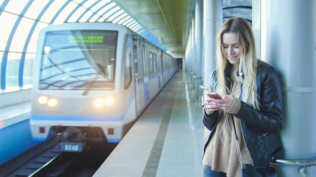 Young girl with gadget long blonde hair in leather jacket with straightens hair standing in metro against the background of a train coming, perspective, horizontal