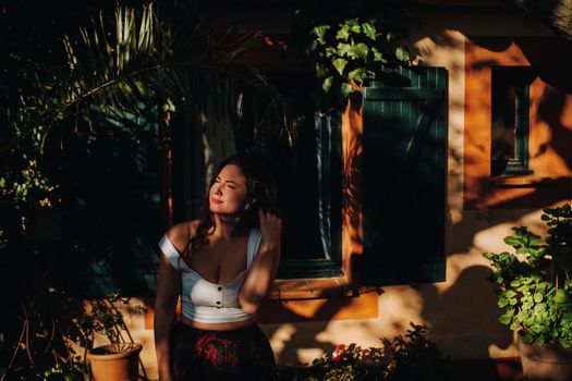 a girl in a white t-shirt with her hair down stands near the window of an old house in France.The girl is smiling, sitting on the window in the skirt of the French town.Provence.