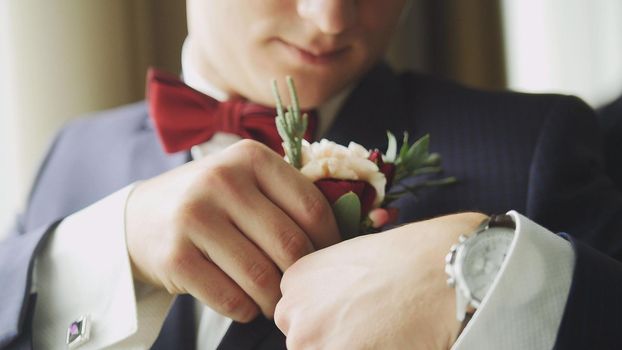 Fashion young man in bow tie and clock - groom's hand arranging boutonniere flower on suit, close up
