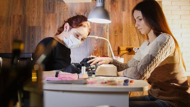 Nail master in medical mask doing professional manicure for white caucasian model, cosmetic salon, telephoto