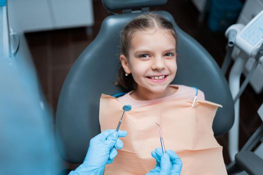 Little cute smiling girl is sitting in dental chair in clinic, office. Woman doctor is preparing for examination of child teeth. Visiting dentist with children.
