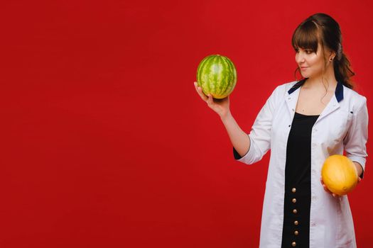 a female doctor nurse in a white coat with fruit in her hands poses on a red background, melon, watermelon