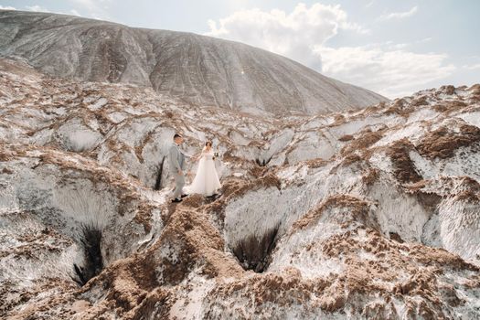 A beautiful couple of lovers posing in a white salt mountain. A young woman in a stylish wedding dress and a beautiful stylish man in a gray suit. The concept of the wedding day