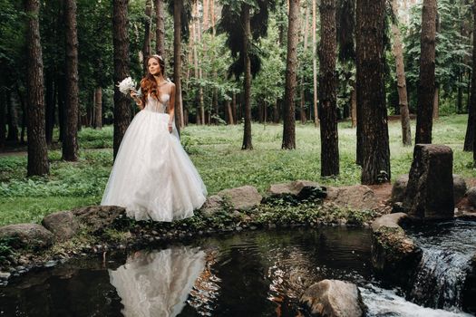 An elegant bride in a white dress and gloves holding a bouquet stands by a stream in the forest, enjoying nature.A model in a wedding dress and gloves in a nature Park.Belarus.