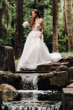 An elegant bride in a white dress and gloves holding a bouquet stands by a stream in the forest, enjoying nature.A model in a wedding dress and gloves in a nature Park.Belarus.