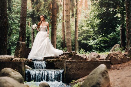 An elegant bride in a white dress and gloves holding a bouquet stands by a stream in the forest, enjoying nature.A model in a wedding dress and gloves in a nature Park.Belarus.