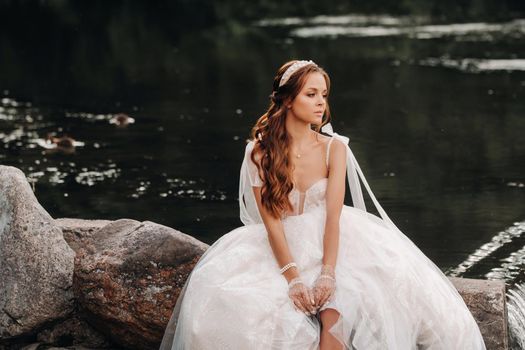 An elegant bride in a white dress and gloves is sitting by the lake in the Park, enjoying nature.A model in a wedding dress and gloves in a nature Park.Belarus