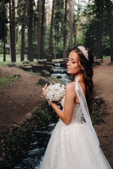 An elegant bride in a white dress and gloves holding a bouquet stands by a stream in the forest, enjoying nature.A model in a wedding dress and gloves in a nature Park.Belarus.