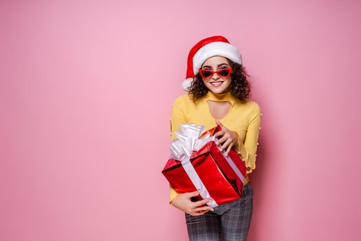 Young happy curly girl in Santa's hat and red sunglasses holds red giftbox while stands on pink background. New Year concept