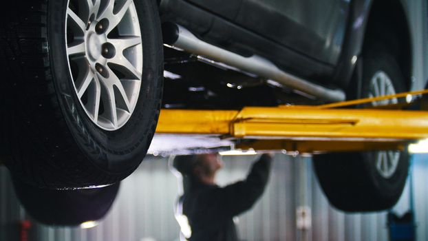 Car lifted in automobile service for fixing, worker repairs detail, telephoto