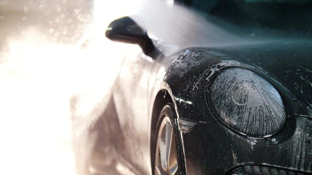 Worker in auto service is washing a luxury car by water hoses, backlight, telephoto