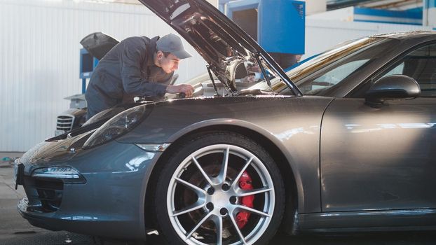 Mechanic male in automobile garage checking hood of the luxury sportcar, telephoto, horizontal