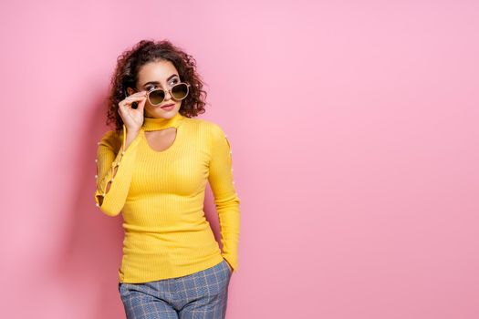 Studio shot of pretty curly caucasian girl wearing in yellow outfit and sunglasses, model posing isolated over pink studio background. Looking away