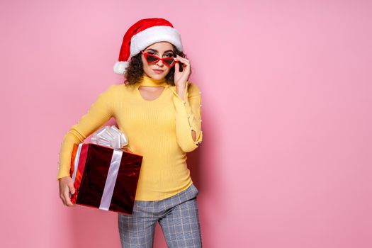 Young curly girl in Santa's hat and red sunglasses holds red giftbox while stands on pink background. New Year concept