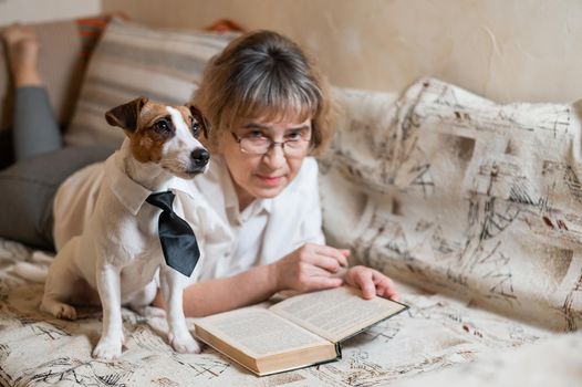 An elderly caucasian woman is lying on a sofa with a smart dog jack russell terrier wearing glasses and a tie and reading a book