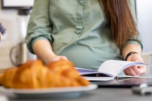 pregnant woman reading book faceless portrait. the concept of women's education, healthy lunch, a gluten-free diet. delicious croissants lie on a plate on the kitchen table in blur.