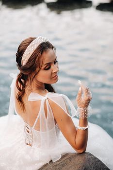 An elegant bride in a white dress and gloves is sitting by the lake in the Park, enjoying nature.A model in a wedding dress and gloves in a nature Park.Belarus