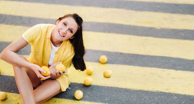 a girl holding lemons in her hands dressed in a yellow shirt, shorts and sitting on a yellow pedestrian crossing in the city and smiling. The lemon mood.