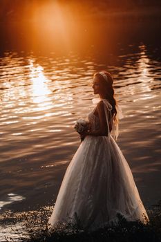 An elegant bride in a white dress and gloves stands by the river in the Park with a bouquet, enjoying nature at sunset.A model in a wedding dress and gloves in a nature Park.Belarus.