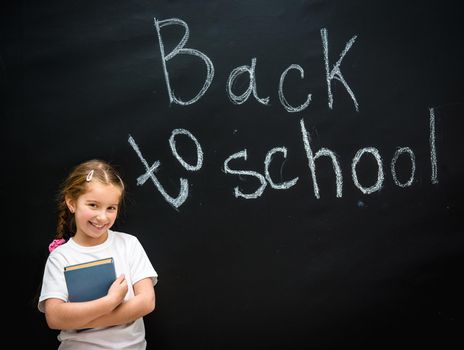 cute little girl with a blue book in hands smiling on black background blackboard on which the back to school