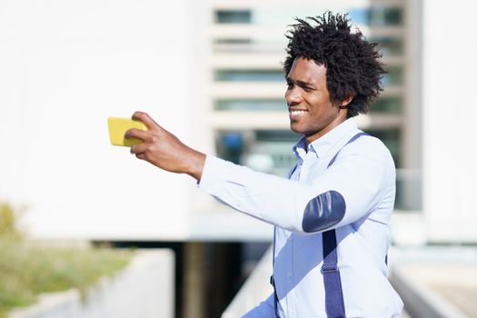 Black businessman with afro hairstyle taking a selfie with his smartphone near an office building.