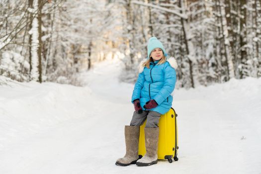 A girl in winter in felt boots sits on a suitcase on a frosty snowy day.